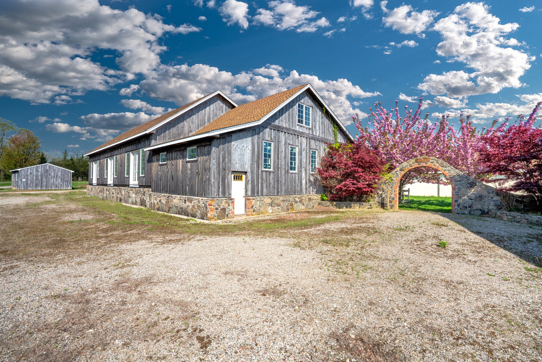 Building 3 in spring — gray weathered wood barn beside the property's stone wall and brick arch with cherry blossoms